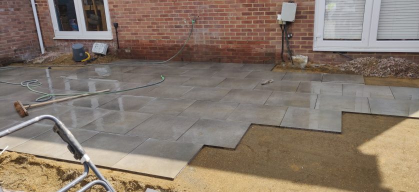 Freshly poured concrete patio with Porcelain tiles, partially completed next to a brick wall.