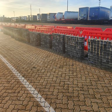 Rows of orange barriers along a brick-paved road near parked trucks.